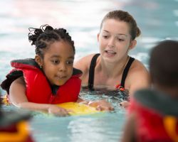 A child in a life jacket swims on a paddle board with an adult and other kids nearby.