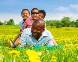 A smiling man lies on his stomach in a field of dandelions. Three smiling kids sit on his back.