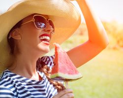 A smiling woman in a hat and sunglasses holding a slice of watermelon on a stick.