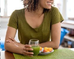A woman holds a glass of green liquid in front of a plate of fruit.