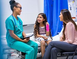 A child with a cast on her arm sits in an exam room with a parent and a doctor.