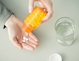 Closeup of a pair of hands spilling pills from a prescription bottle into one palm. A glass of water sits to the side.