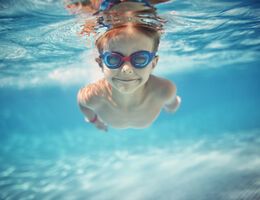 A boy in goggles smiles as he swims under the water in a pool.