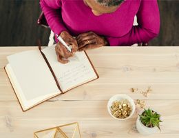 A woman sits at a table writing in a journal.