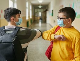 Two masked grade school kids touching elbows in a school hallway.
