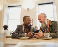 Two men in a coffee shop look at a smartphone and smile at each other.