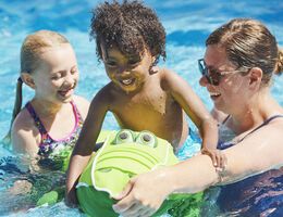 A woman helps a boy balance on a pool toy as a girl looks on.