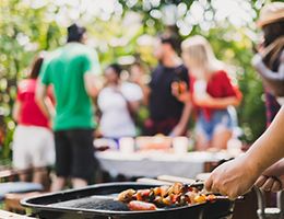 A person puts kebabs on a grill with partygoers in the background.