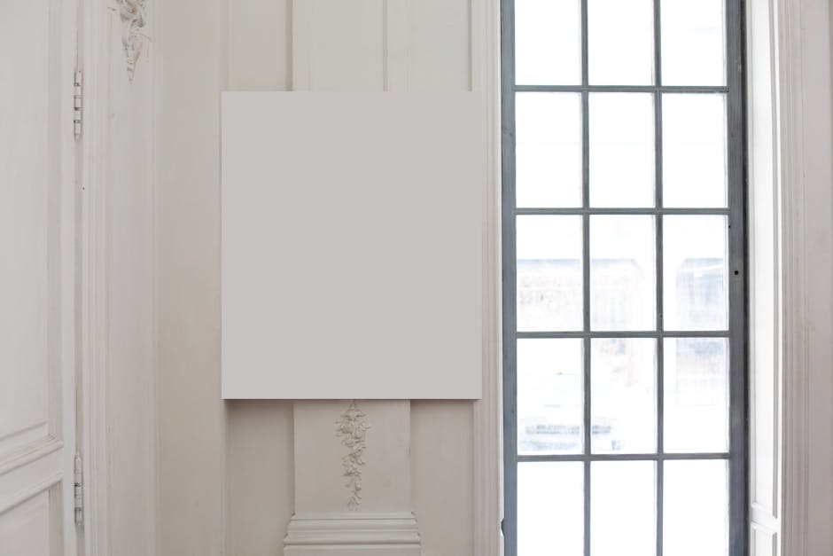 A blank white canvas hangs on a wall beside a large, grid-patterned window in a minimalist, light-filled room.