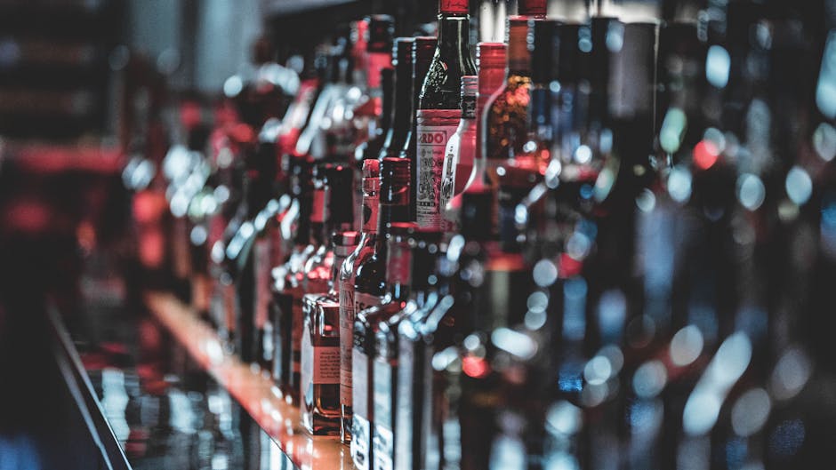 A close-up shot of various liquor bottles lined up on a bar counter. The image captures a wide assortment of bottles, each with different shapes and labels, suggesting a diverse selection of alcoholic beverages available at the bar. The background is slightly out of focus, emphasizing the collection of bottles in the foreground.