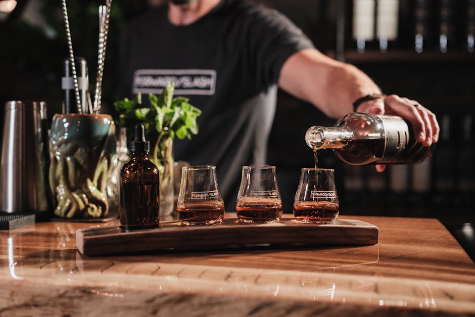 A bartender pouring liquor into glasses at a wooden bar. The scene includes three glasses with alcohol, a brown bottle, and a decorative mug with bar tools and herbs in the background.