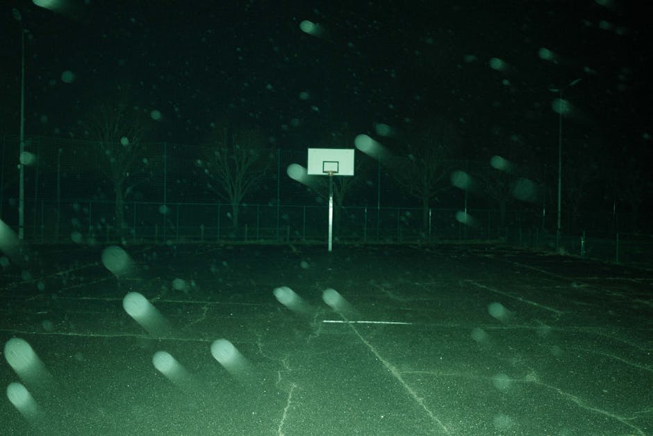 A dimly lit outdoor basketball court at night with a single hoop in the center, captured under snowfall or rain.