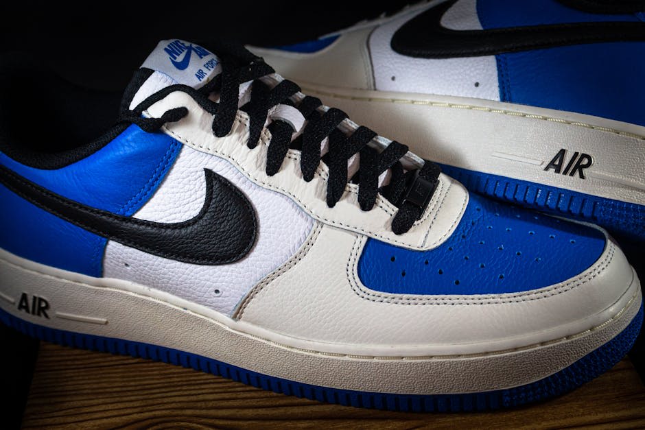 Close-up image of a pair of low-top Nike sneakers with a blue and white color scheme, featuring the iconic black swoosh logo on the sides. The shoes have 'AIR' written in black on the white sole, highlighting their brand and style. The laces are black, and the sneakers are positioned on a wooden surface, showcasing their design and details.