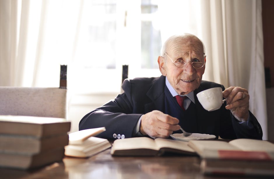 An elderly man in a suit sits at a table with books, holding a cup of tea or coffee inside a brightly lit room.