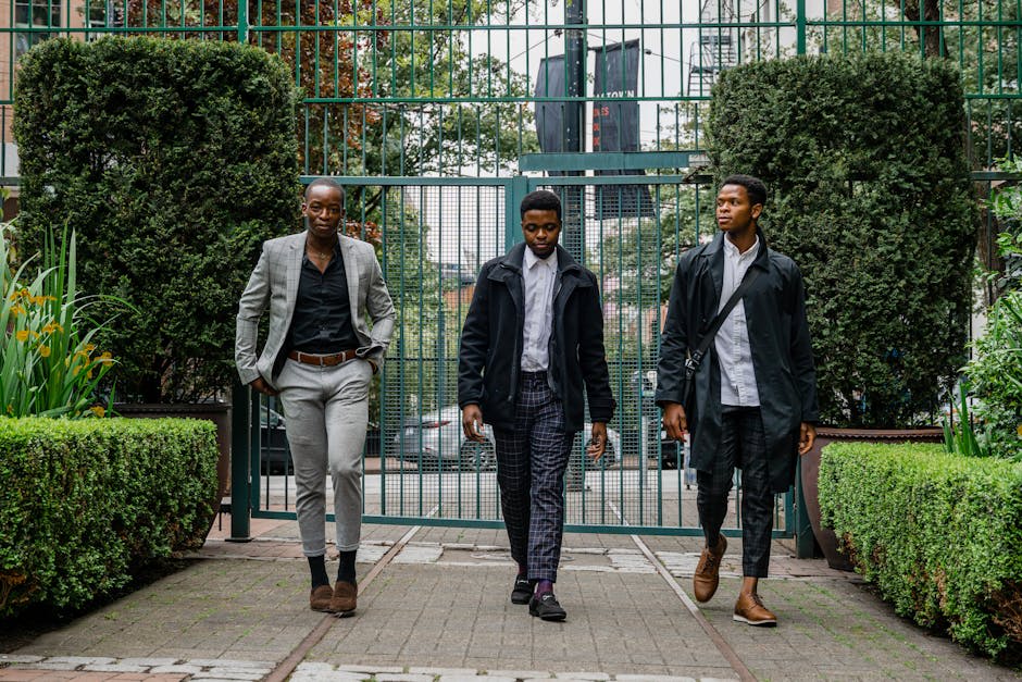 Three men in formal and semi-formal attire walk side by side in an outdoor garden with a gated entrance in the background.