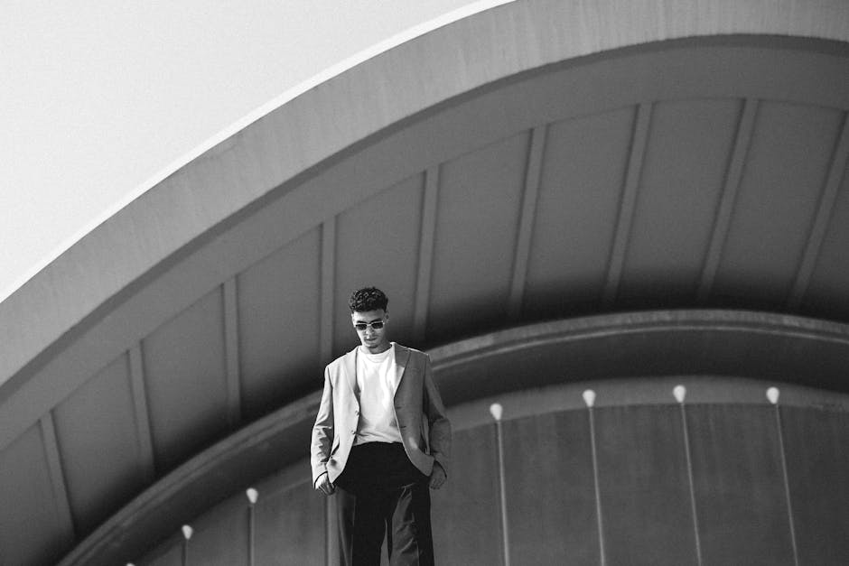 A black-and-white image of a man in sunglasses and a blazer standing in front of an arched, large architectural structure.