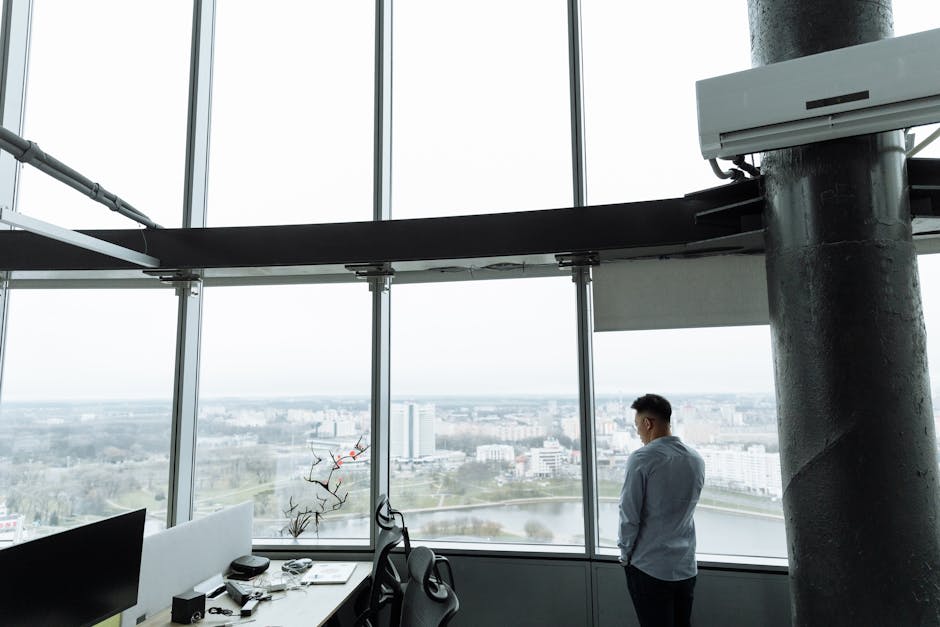 A man standing by large floor-to-ceiling windows in a modern office, gazing out at a cityscape.