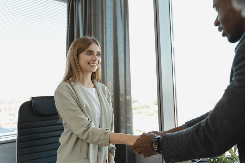 A woman and a man engaged in a handshake in an office setting. The woman is smiling and appears pleased, with a view of the city through the large window behind them. The setting suggests a professional business meeting or interview.