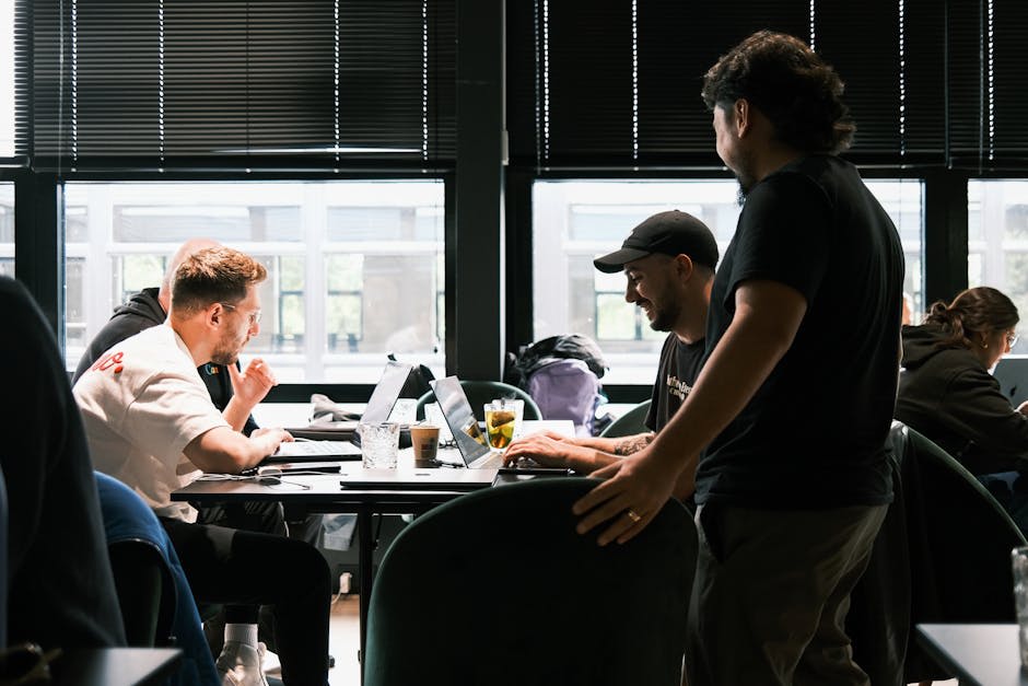 Three people are collaborating and working on laptops at a table in a dimly lit modern office setting.
