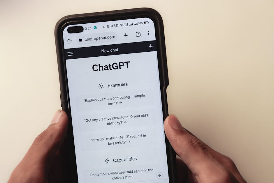 A person holding a smartphone displaying the ChatGPT interface on a web browser. The screen shows options such as 'Explain quantum computing in simple terms', 'Got any creative ideas for a 10 year old's birthday?', and 'How do I make an HTTP request in Javascript?'. The hand is holding the phone against a plain background.