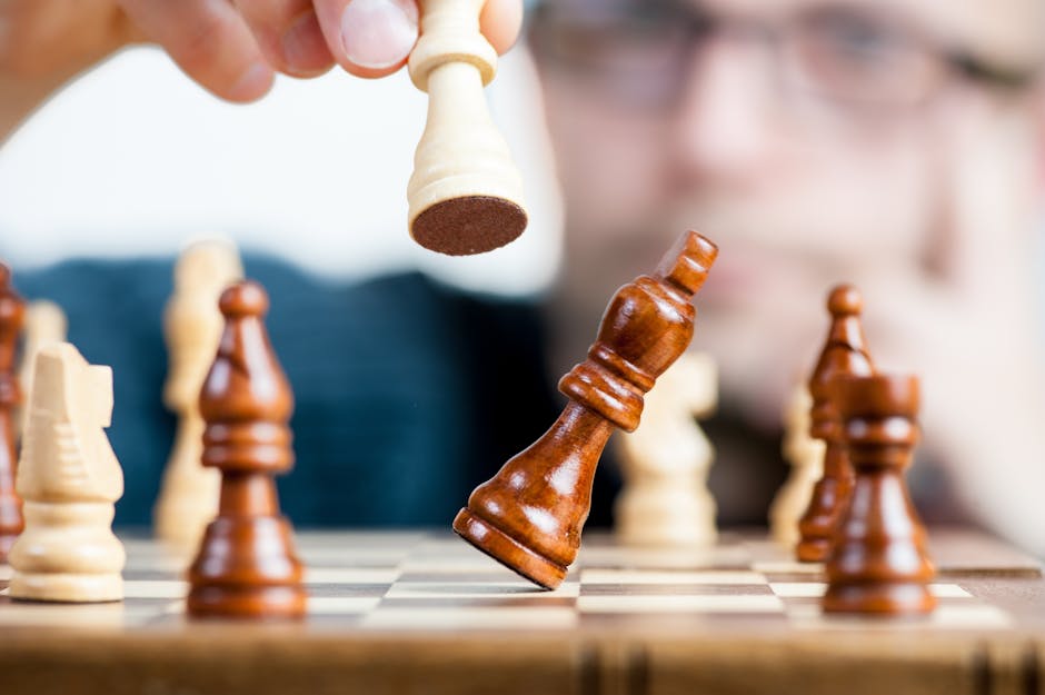 Close-up image of a chess game in progress. A person's hand is about to make a move with a white rook that captures a black king on the chessboard. The focus is on the chess pieces, showcasing action and strategy, with blurred background emphasizing concentration.