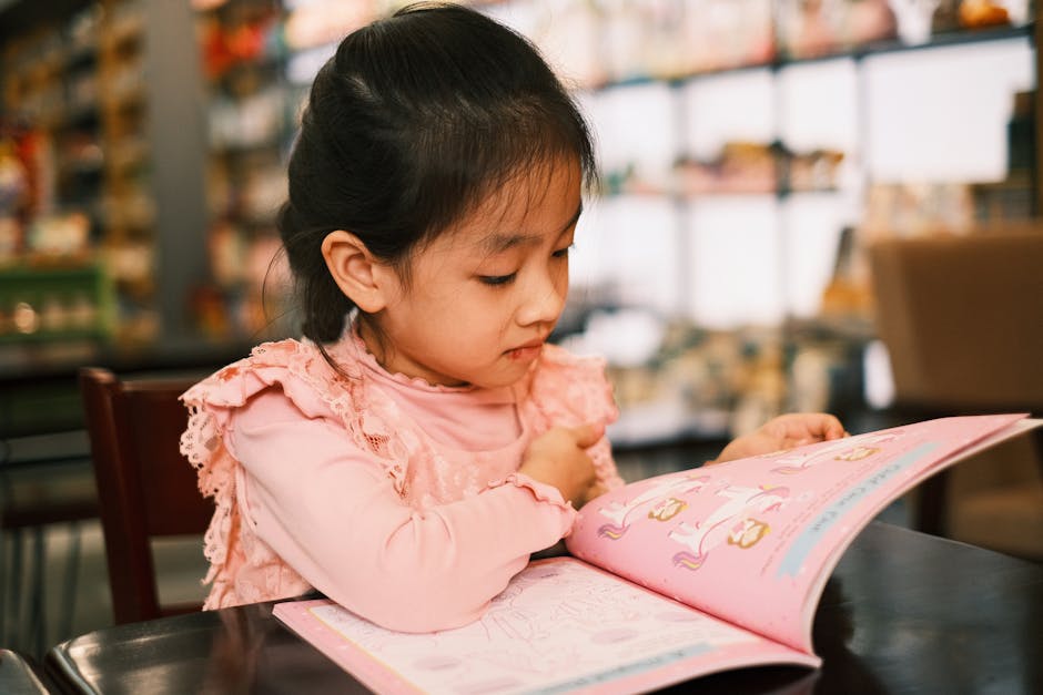 A young girl in a pink outfit is sitting at a table reading a book attentively. The background shows a bookstore setting with shelves filled with various items. The focus is on the child's engagement with the book.