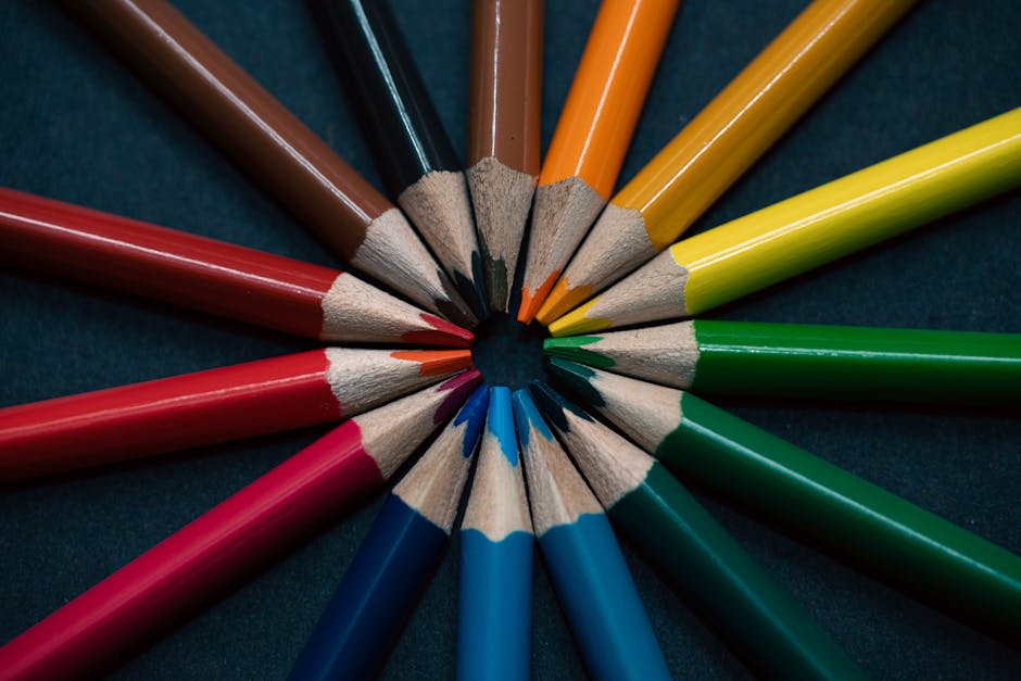 A close-up image of several colored pencils arranged in a circular pattern with their tips pointing towards the center on a dark background. The pencils are vibrant and include colors such as red, blue, green, yellow, orange, and brown. This composition highlights art supplies and creativity.
