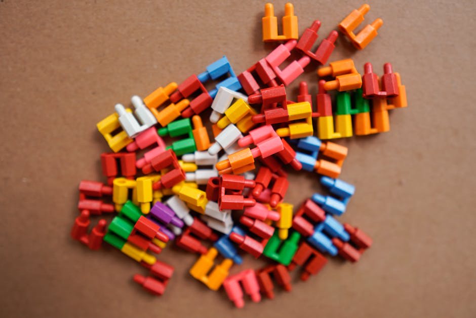 A pile of colorful plastic game pieces on a brown cardboard surface. The pieces are small, two-pronged, and come in various colors including red, yellow, orange, blue, green, and white. These are likely used in a board game or a creative building activity.