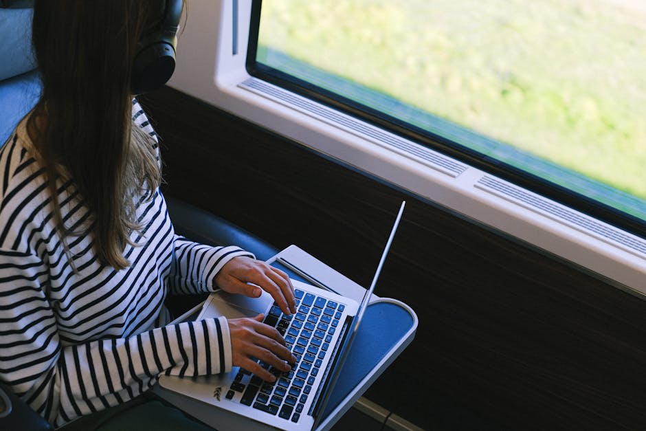 A person wearing headphones works on a laptop while seated next to a window on a train.
