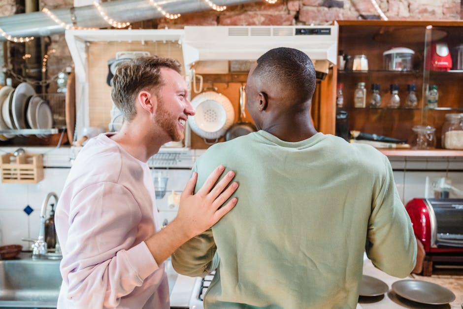 Two people are standing together in a kitchen, with one smiling and placing a hand on the other's shoulder.