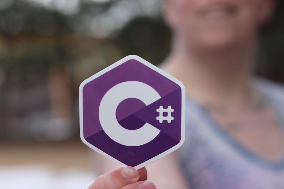 A close-up image depicting a person holding a sticker featuring the C# programming language logo. The logo is hexagonal with a purple background and a white letter 'C' and a hashtag (#) symbol. The person in the background is blurred, focusing the attention on the sticker in the foreground.