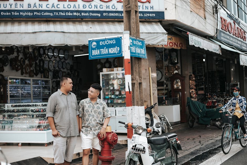 Two people stand near a fire hydrant and street signs at an intersection in an urban area, with a bicycle passing by and shops in the background.