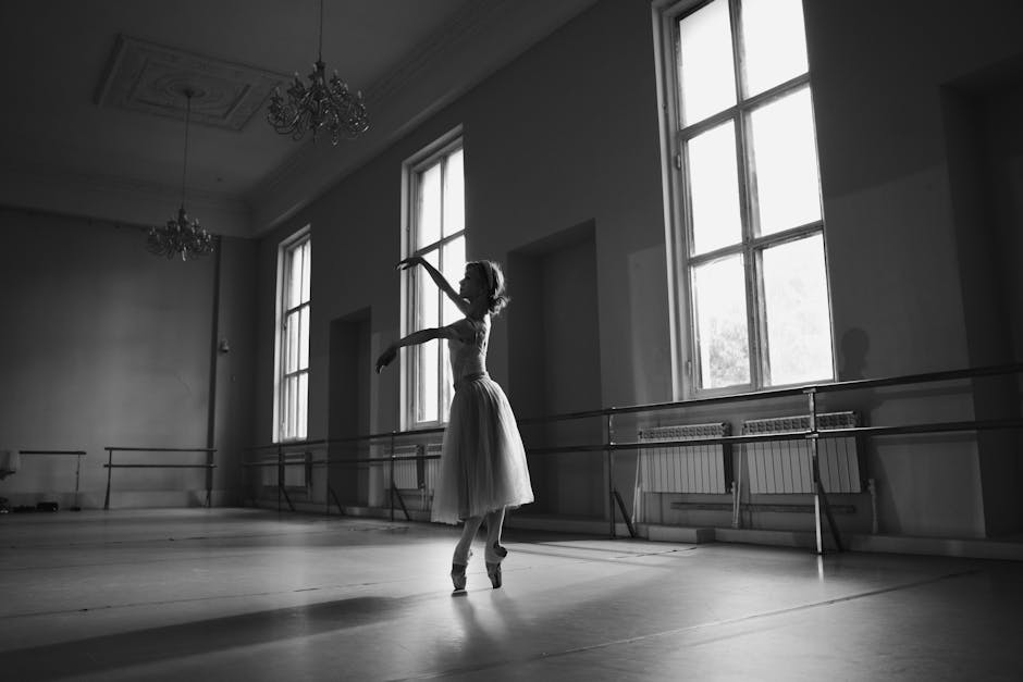 A ballerina practices in an empty dance studio, captured in black and white against large windows and mirrored walls.