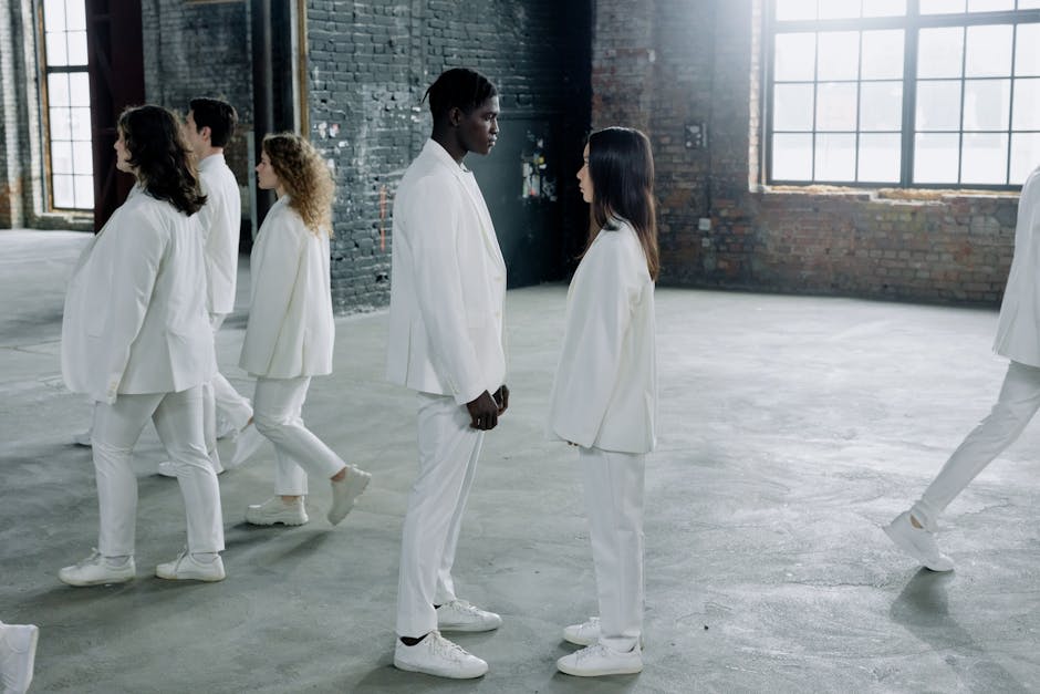 A group of people dressed in white suits stand in a spacious, industrial setting, with two individuals facing each other in the center.