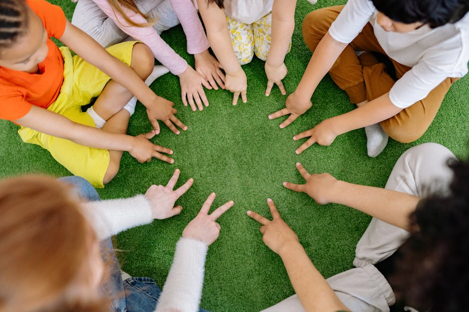 A group of children sit in a circle on grass, extending their hands towards the center as they make peace signs.