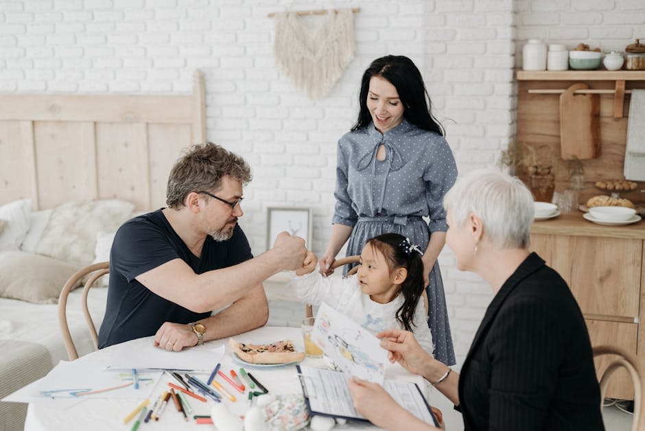 A family gathering around a table, engaged in an art activity. A man and a young girl are fist-bumping, while a woman watches on with a smile. An older woman, likely the girl's grandmother, is holding a drawing. The setting appears to be a cozy, well-lit kitchen or dining area with art and crafts supplies on the table.