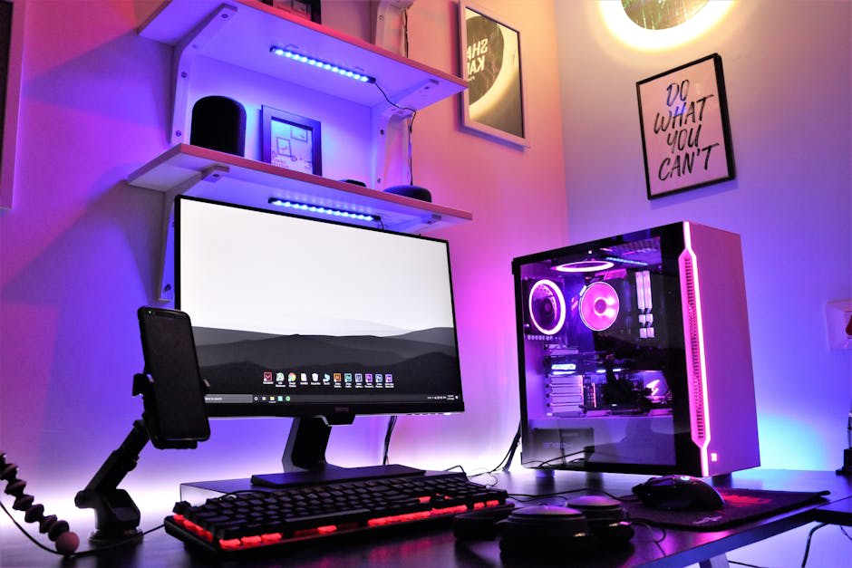 A neatly organized gaming and workstation setup featuring a desktop PC with RGB lighting, a large monitor, keyboard, mouse, and smartphone stand, surrounded by shelves and motivational wall art.