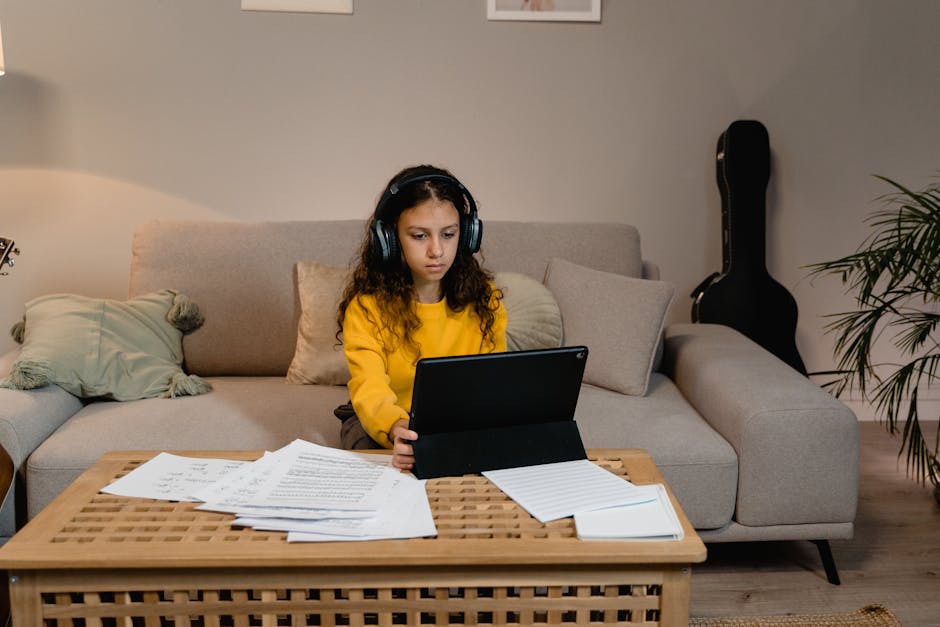 A young girl in a yellow sweater is sitting on a beige sofa, wearing headphones and using a tablet. She is focused on the device in front of her. In the foreground, there is a wooden coffee table with scattered papers. The living room setting includes a guitar case leaning against the wall and some decorative cushions.