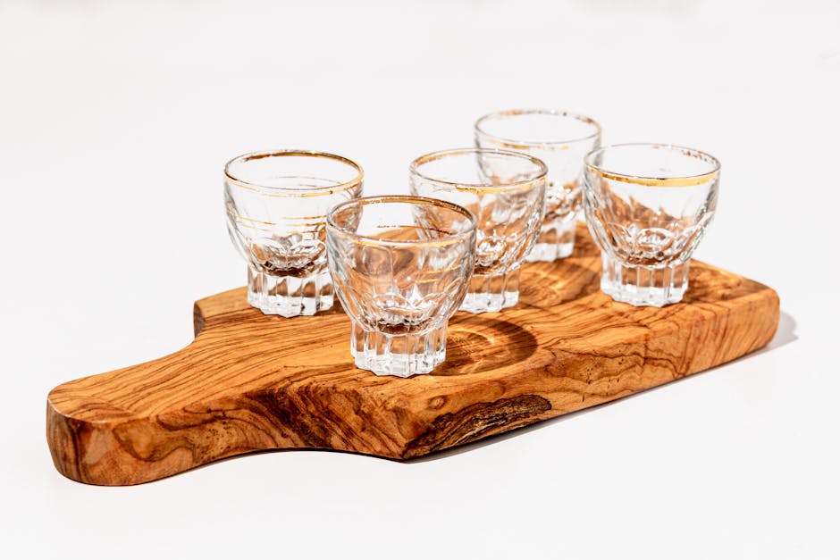 The image depicts five clear glass shot glasses placed on a wooden serving board. The board has a natural wood grain pattern and is set against a light background. The shot glasses are empty and arranged neatly, showcasing their intricate designs.