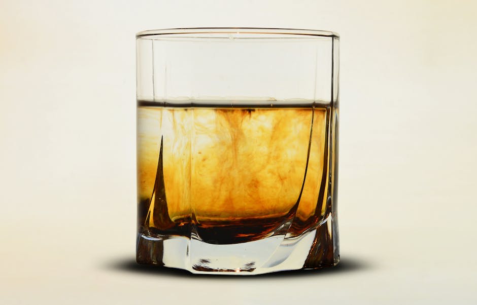 A clear glass tumbler approximately half-filled with amber-colored whiskey. The whiskey has a swirling effect inside, giving it a rich texture against the light background. The glass has a simple, modern design with light reflections on its edges.
