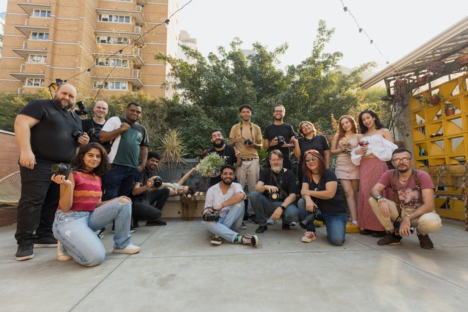A large group of photographers posing together outdoors. The group consists of diverse individuals holding cameras, suggesting they are either participating in a photography event or workshop. The setting appears to be a patio or outdoor area with buildings and greenery in the background, creating a casual and relaxed atmosphere. The image captures a sense of community and shared interest in photography.