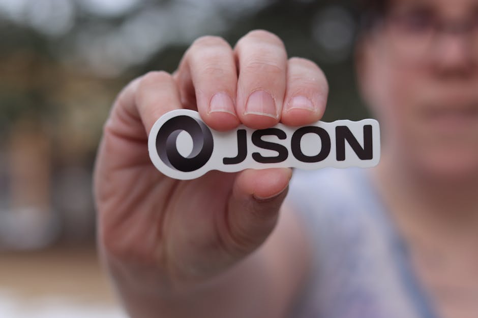 A close-up image of a person's hand holding a sticker with the word 'JSON' on it. The background is blurred, focusing attention on the sticker. Useful for themes related to programming, data formats, or technology.