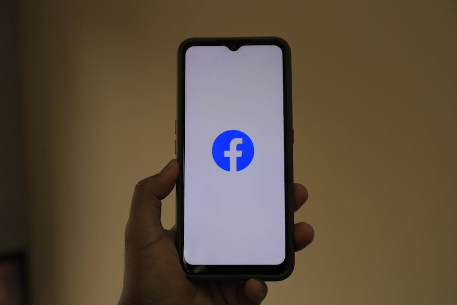 A hand holding a smartphone displaying the Facebook logo on its screen. The phone is centered against a plain background, focusing on the use of social media.