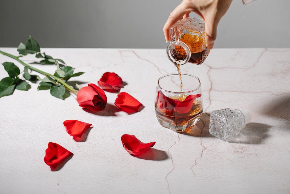 A hand pouring whiskey or a similar beverage into a glass that contains rose petals. The scene is set on a marbled surface with scattered red rose petals and a long-stemmed red rose lying nearby. An empty crystal bottle rests on its side.