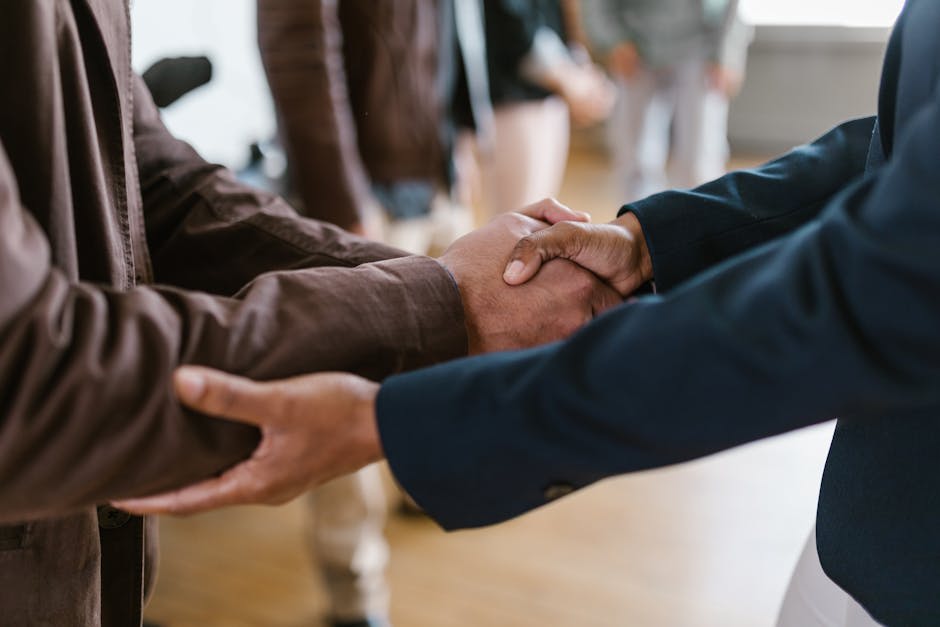 A close-up image depicting a handshake between two individuals, each wearing business attire. The focus is on their hands, symbolizing a professional agreement or partnership. The background is blurred, indicating a formal or business setting.