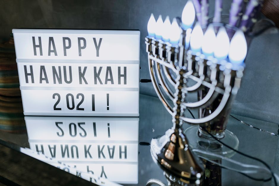 The image shows a 'Happy Hanukkah 2021!' sign with a lit menorah in the background. The menorah is on a reflective surface, enhancing the festive atmosphere. The image captures the essence of the Hanukkah celebration with lit candles.