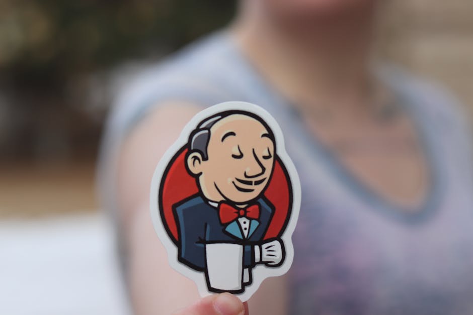 A person holding a sticker featuring the Jenkins logo, depicted as a dapper cartoon butler with a red bowtie and a tuxedo. The focus is on the sticker, with the person blurred in the background, indicating a casual setting.