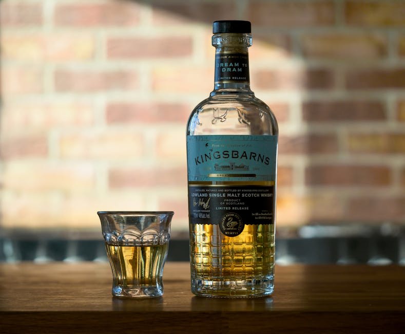 A bottle of Kingsbarns single malt Scotch whisky with a low glass containing a small amount of whisky, both placed on a wooden surface. The background features a blurred brick wall, creating an inviting atmosphere for enjoying whisky.