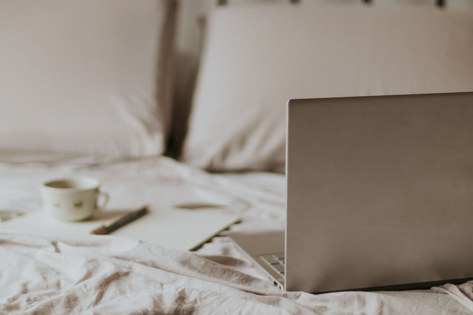 A laptop, cup of coffee, notebook, and pencil are placed on a rumpled bed with pillows in the background.