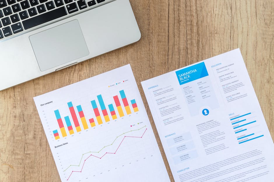 The image shows a wooden desk with a silver laptop partially visible. Next to the laptop, there are two sheets of paper. The first sheet is a bar and line chart with colorful data. The second sheet appears to be a resume with the name 'Samantha Black' at the top. The resume includes sections for experience, education, skills, and references, presented in a clean layout. This image is ideal for representing business analysis or job application themes.